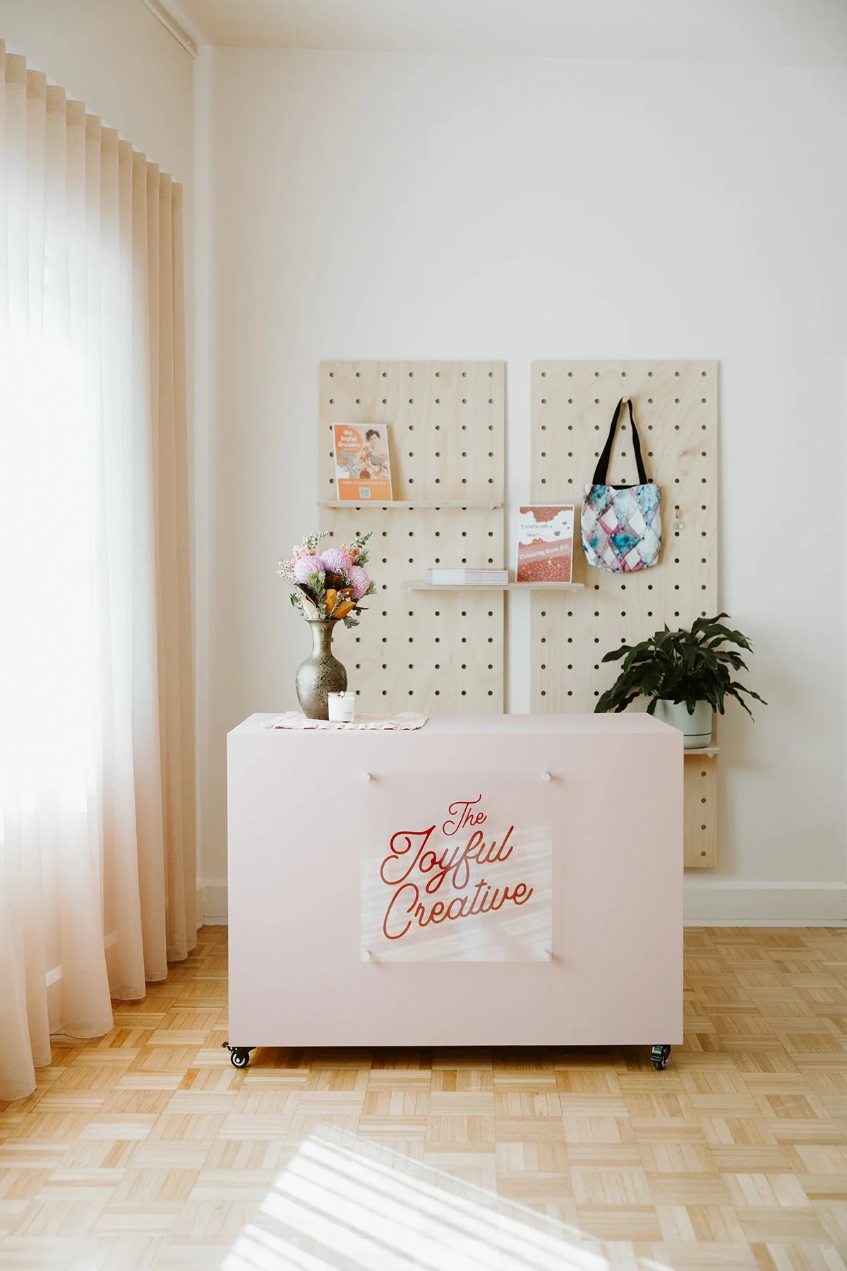 A reception desk with a sign that reads 'The Joyful Creative' in red cursive, a flower vase with pink and white flowers, a small white candle, and a pegboard background with books, a flyer, a tote bag, and a potted plant.