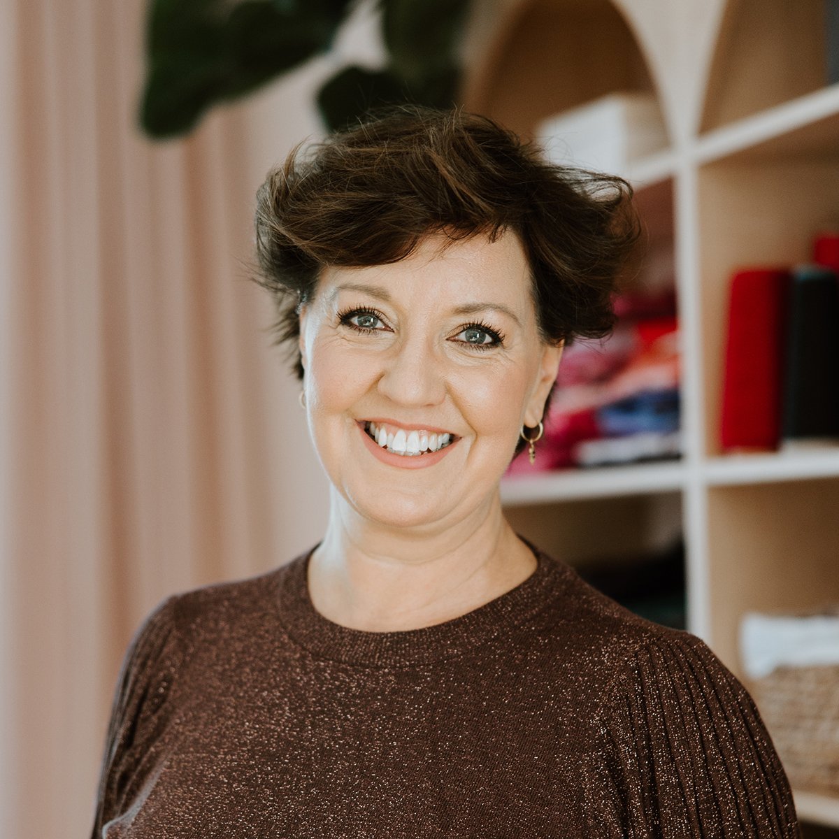A smiling middle-aged woman with short brown hair, wearing a brown sparkly top, standing in a room with shelves and a large plant in the background.