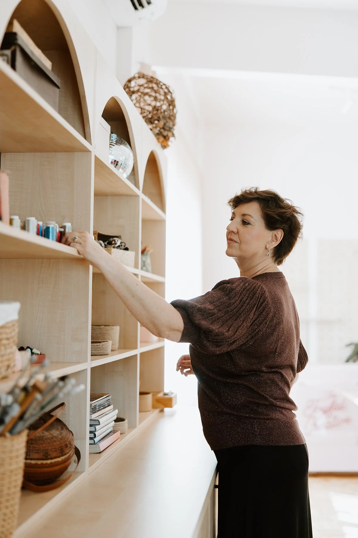 A woman with short brown hair in a brown top reaches for items on a light wood bookshelf filled with books, baskets, and decorative objects.