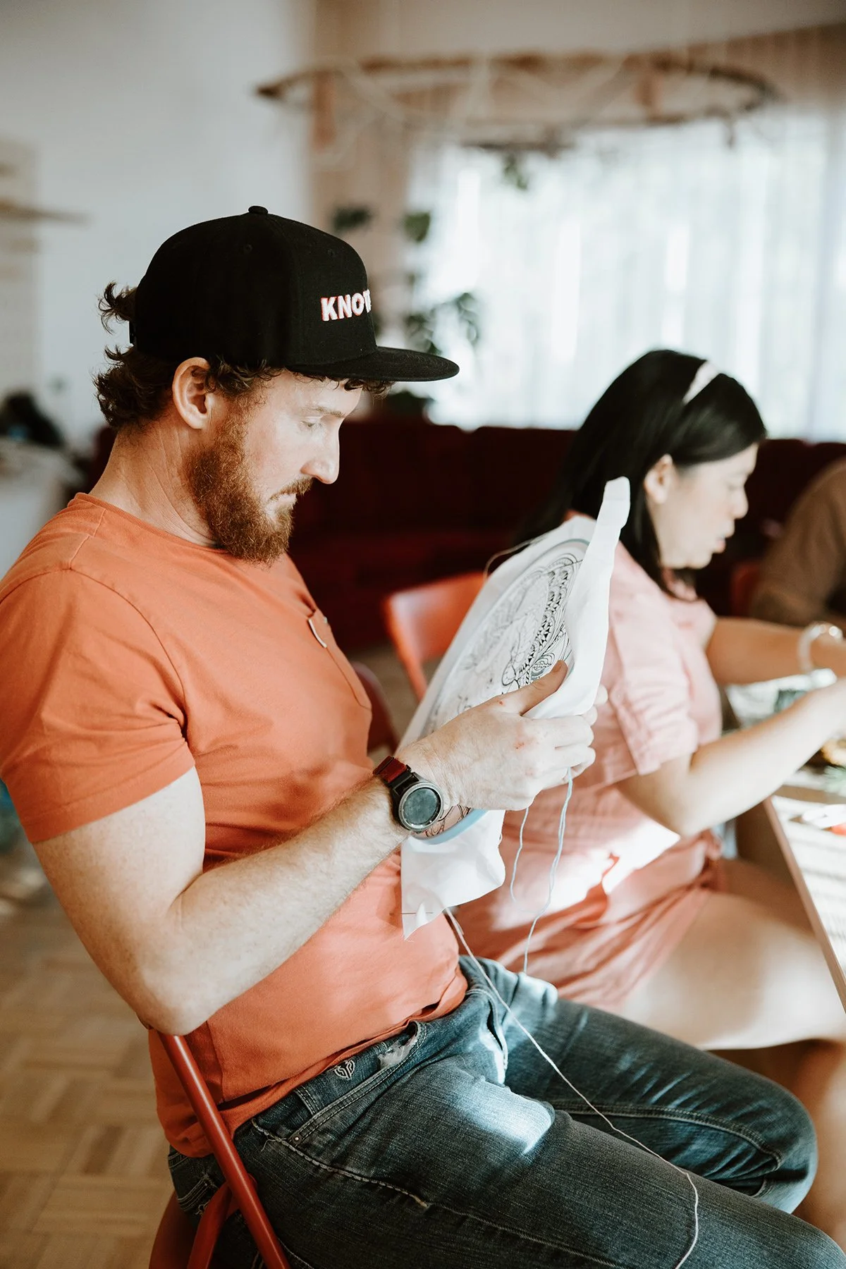 A man with a beard wearing a black cap and orange shirt looking at a coloring book, sitting on a red chair, with another woman next to him also coloring, in a bright room with natural light