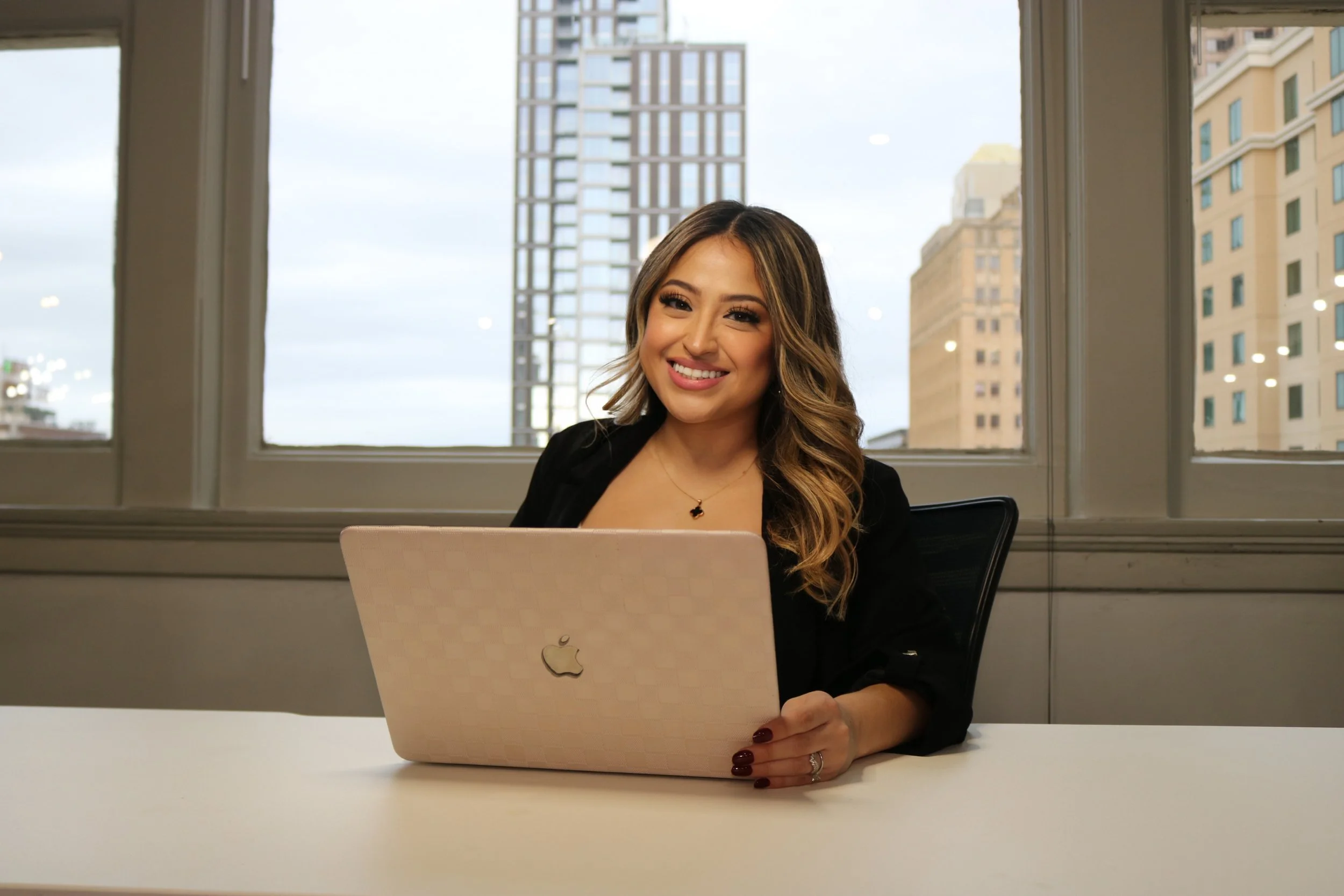A woman with long wavy brown hair sits at a desk with a pink Apple laptop in front of her, smiling at the camera. She is wearing a black blazer and has a cityscape with tall buildings visible through large windows behind her.