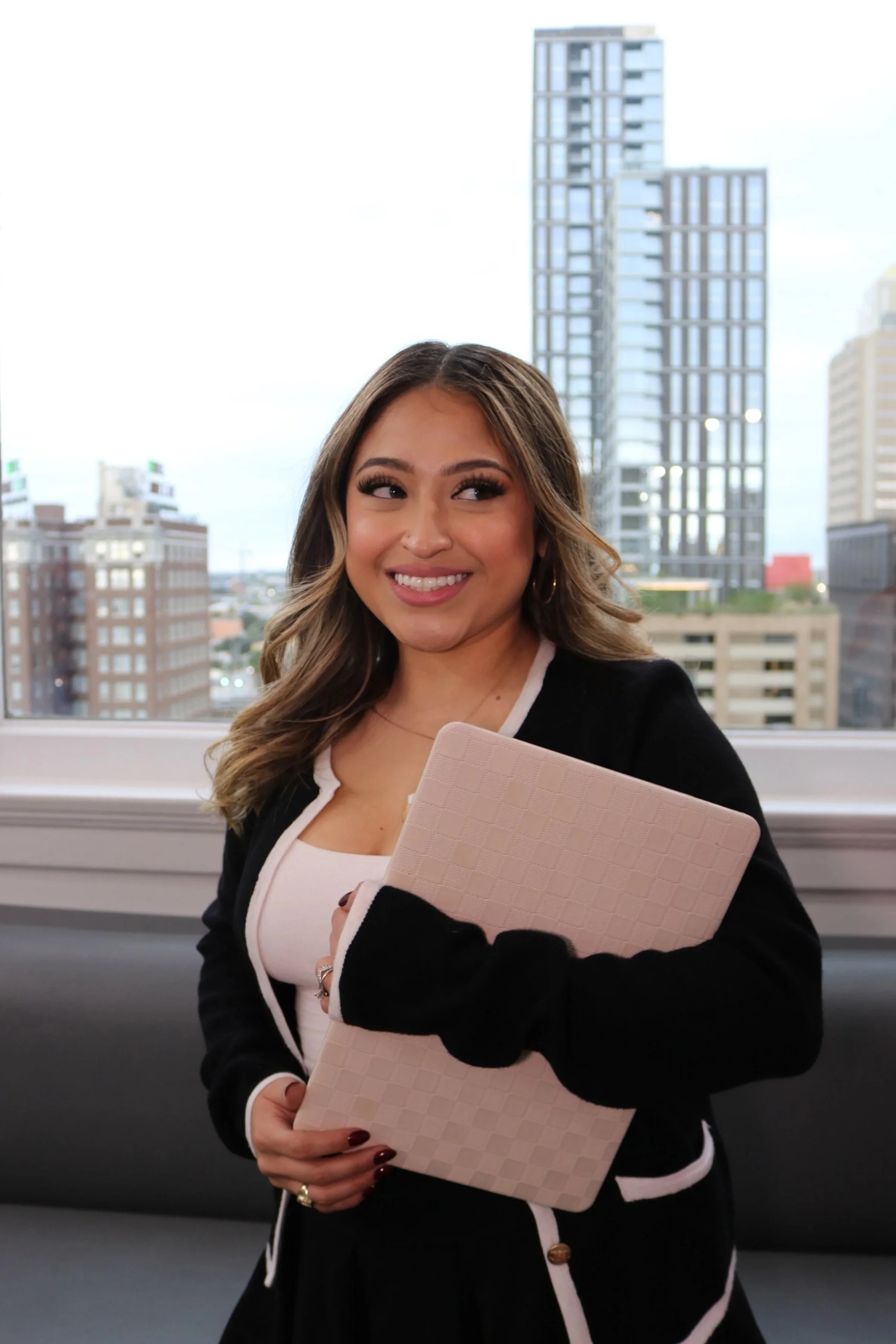 A young woman with wavy hair and hoop earrings smiling while holding a laptop, standing in front of a large window with a cityscape view of tall buildings.