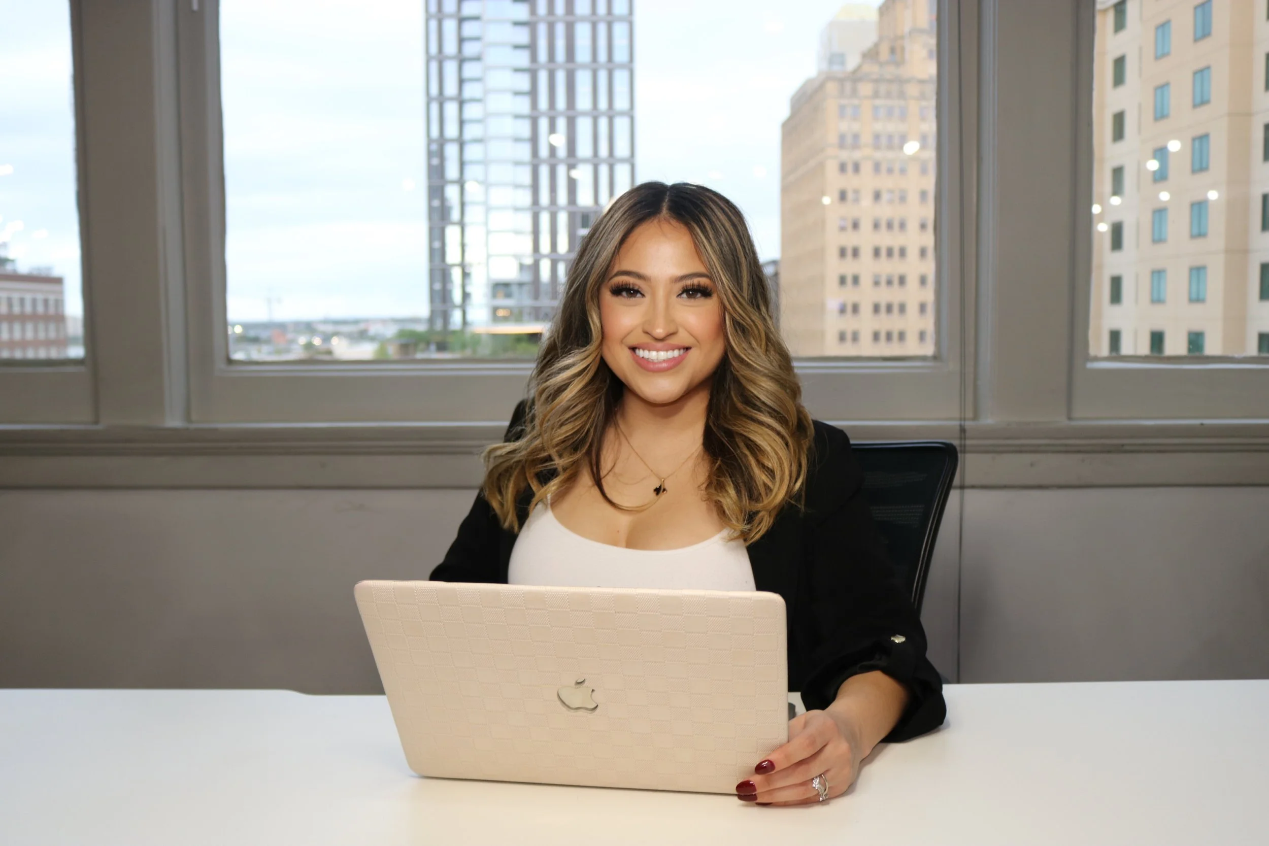 A young woman with wavy blonde hair smiling at a camera, sitting at a white desk with a beige MacBook laptop, in an office with large windows showing a cityscape of tall buildings.