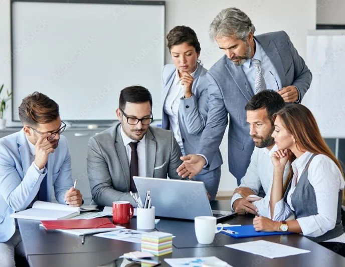 A business team of six professionals collaborates around a laptop in a modern office conference room, with documents and coffee mugs on the table.