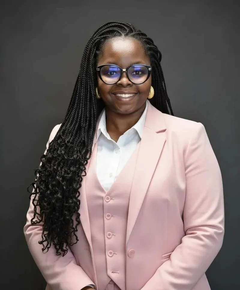 A smiling woman with long braided hair, wearing glasses, gold earrings, a white shirt, and a light pink suit, standing against a dark background.