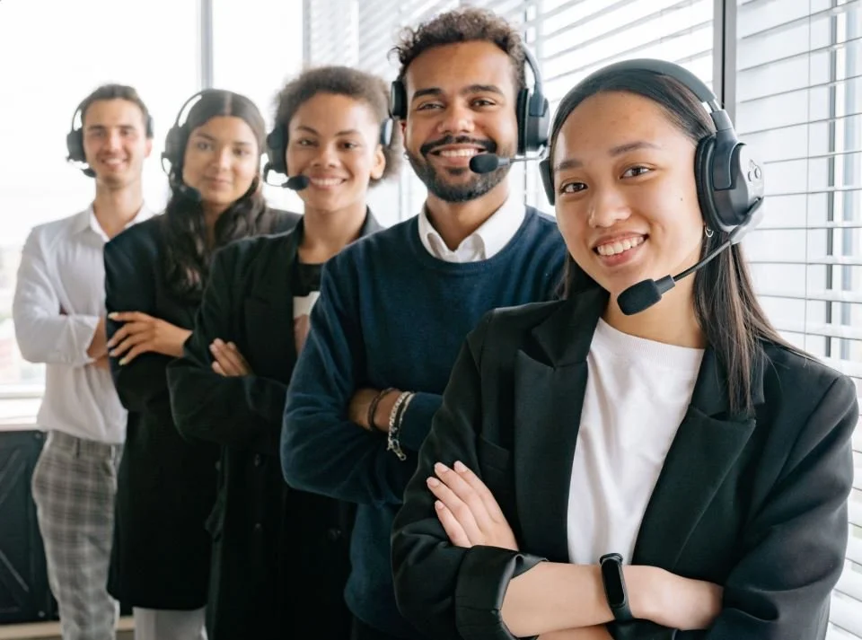 A diverse group of five customer service representatives standing in an office with large windows, wearing headsets and smiling.