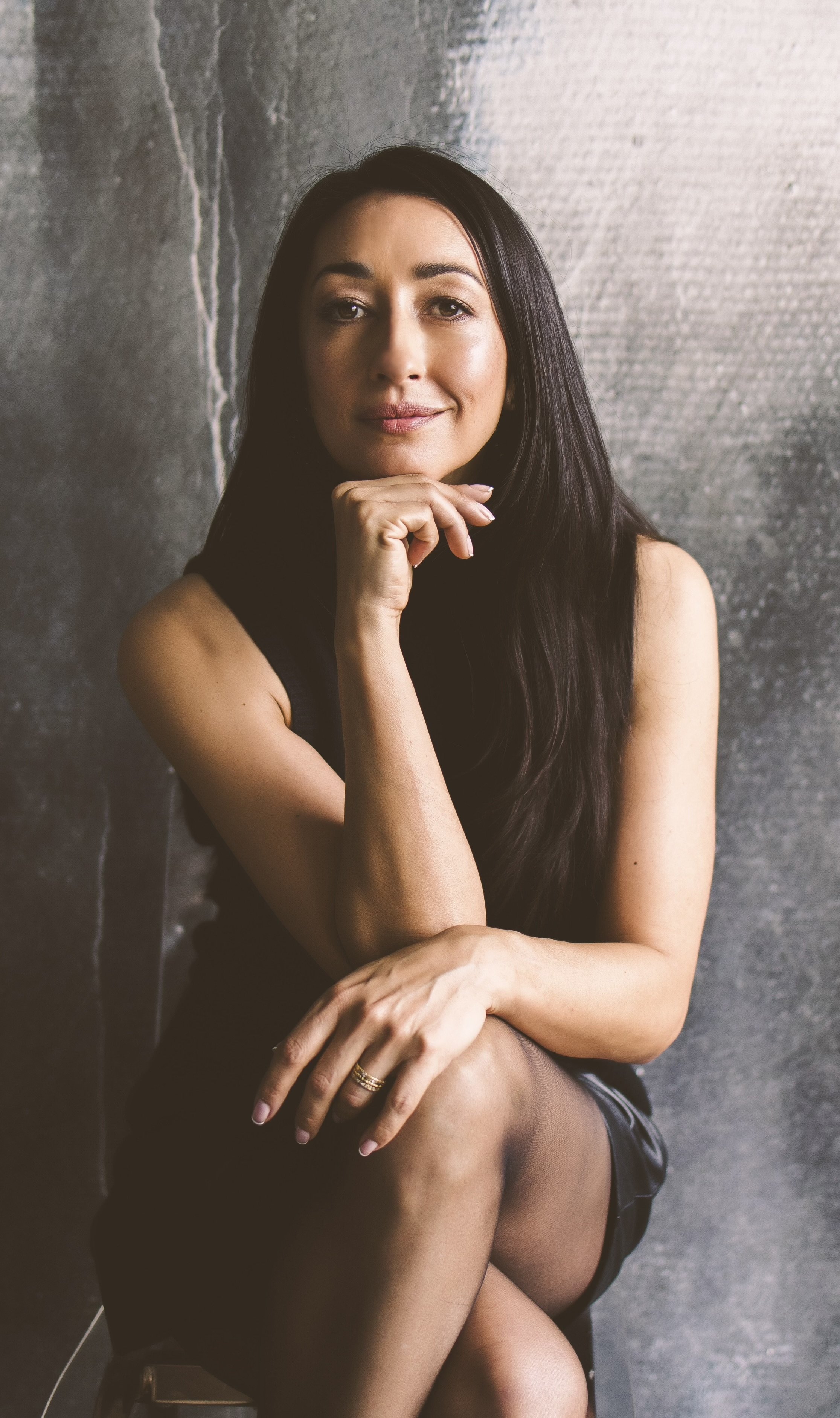 Three-quarter photo portrait of artist Eloise Parker, wearing black, perched cross-legged on a stool in front of a textured wall.