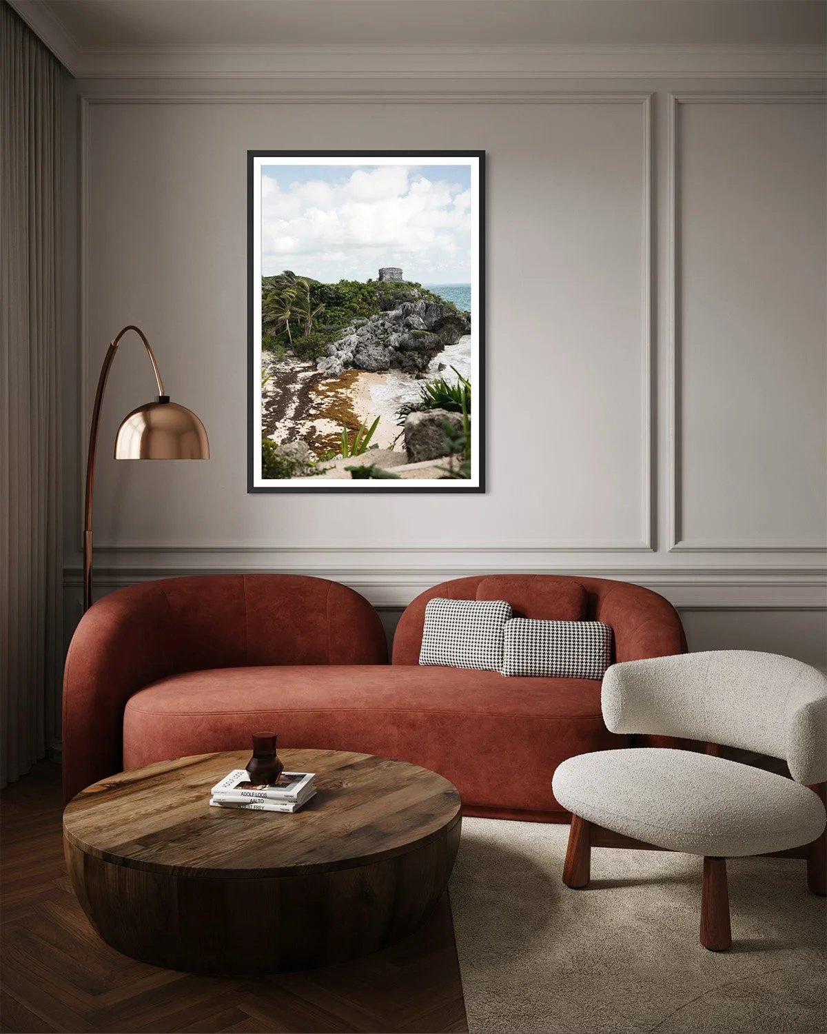 Living room with a coral-colored sofa, a white textured armchair, a wooden coffee table, a gold floor lamp, wall molding, and a framed landscape photograph of a coastal scene with rocks, plants, and a small stone structure.