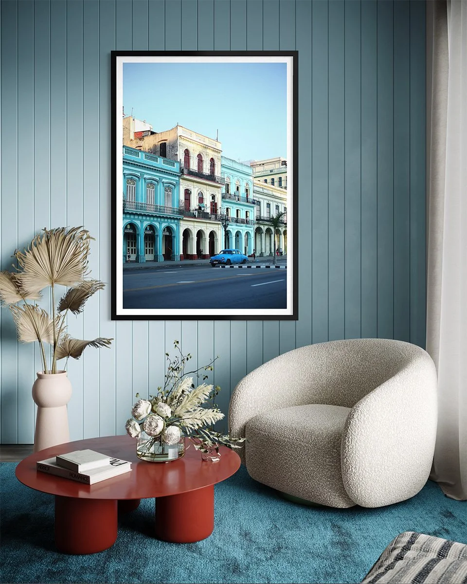 A framed photo of colorful buildings and a street scene hangs on a light blue paneled wall in a cozy living room. The room features a white rounded armchair, a beige vase with dried pampas grass, and a red round coffee table with books and a floral arrangement, with a blue carpet on the floor.
