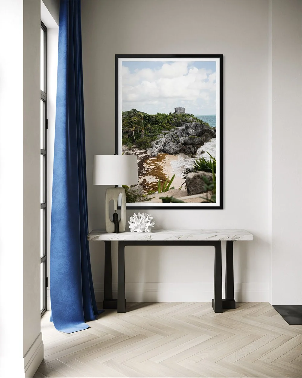 Interior of a room with a large framed beach scene painting, a marble-topped console table, white and black decorative objects, and a window with dark blue curtains.