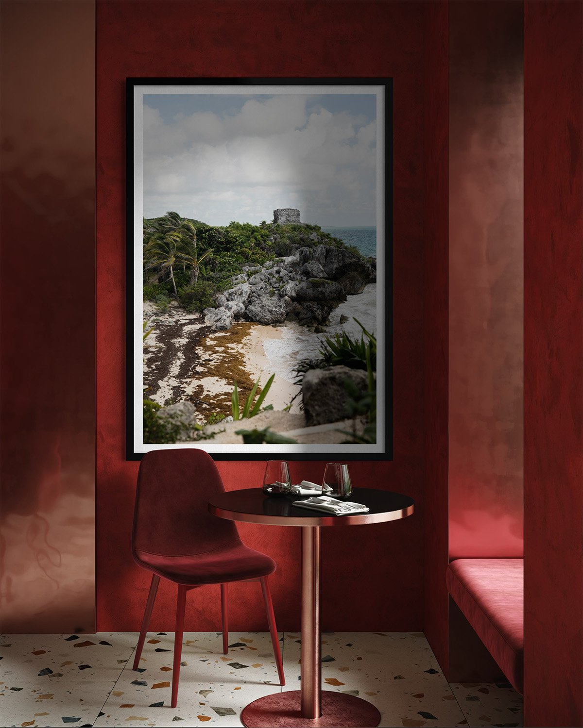 Interior of a restaurant with a small round table set with two glasses, a fork, and a knife, a red upholstered chair, and a large framed photograph of a tropical beach scene with rocks, trees, and an ocean view.