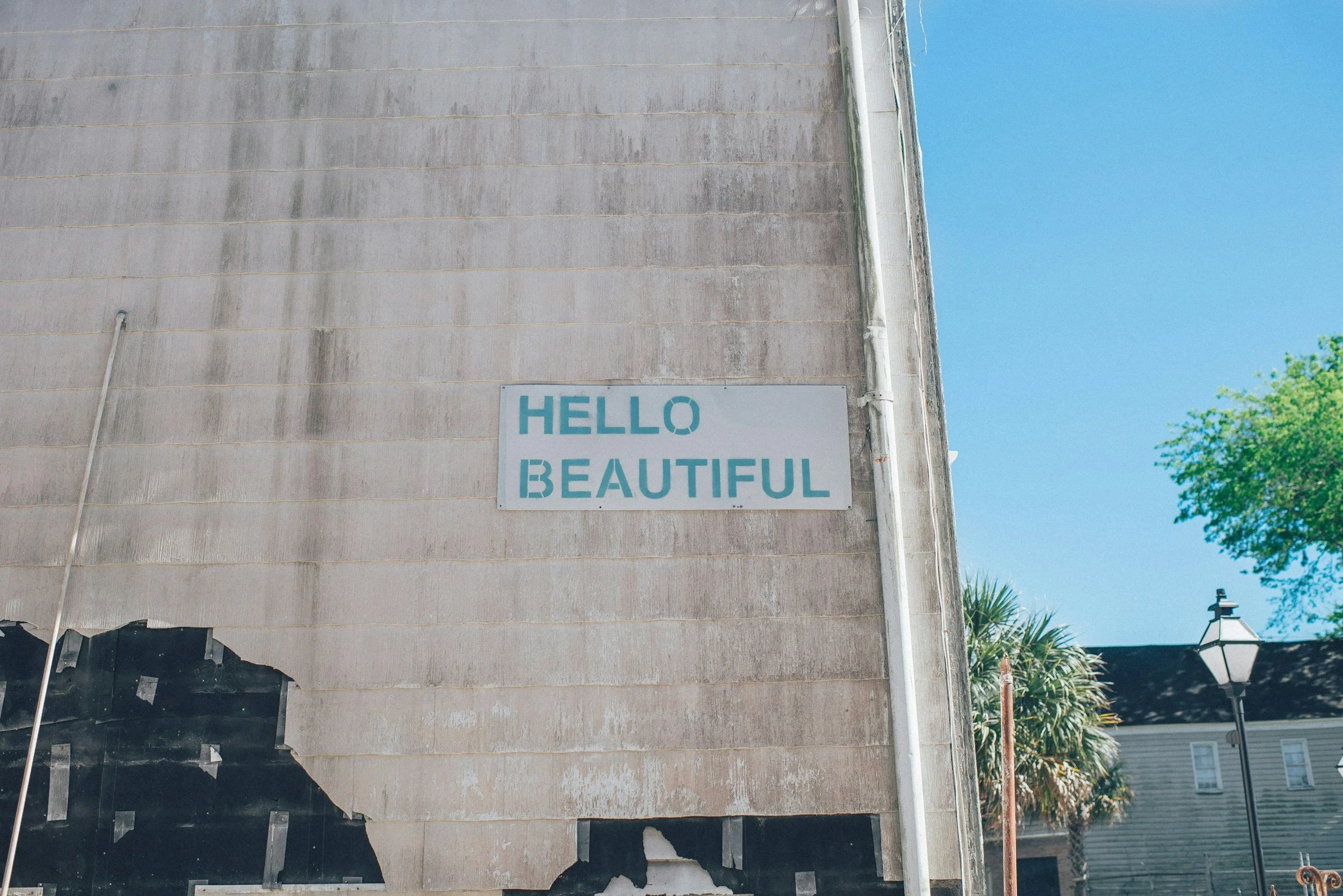 A weathered sign on a beige wall reads 'HELLO BEAUTIFUL' in light blue block letters. There is a blue sky and a green tree in the background.
