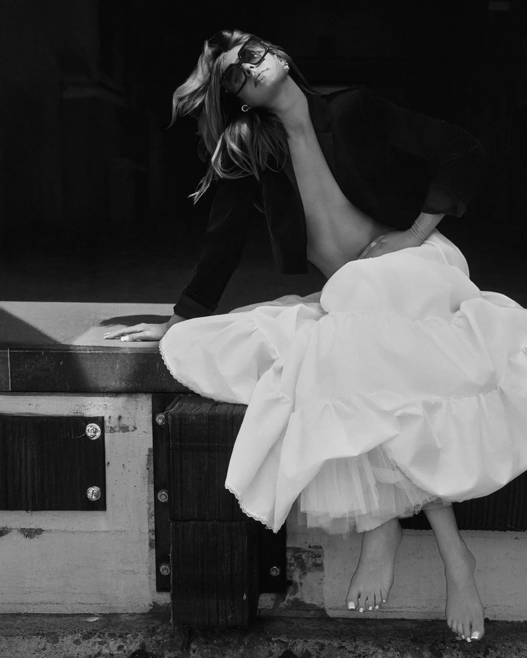 Black and white photograph of a woman sitting on a surface with her legs hanging down, wearing sunglasses, a black jacket, and a long white skirt, with her shirtless upper body partly visible.