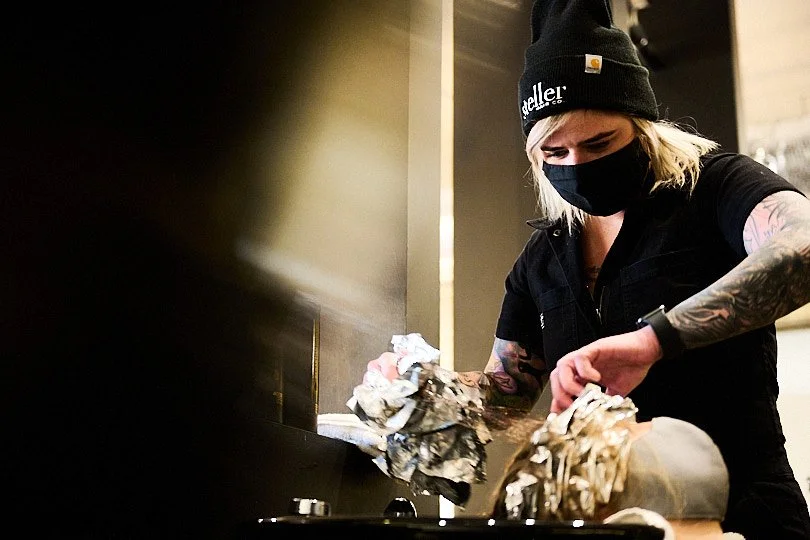 A woman wearing a black beanie and face mask, with tattooed arms, is cleaning or preparing food with foil on a kitchen or restaurant counter.