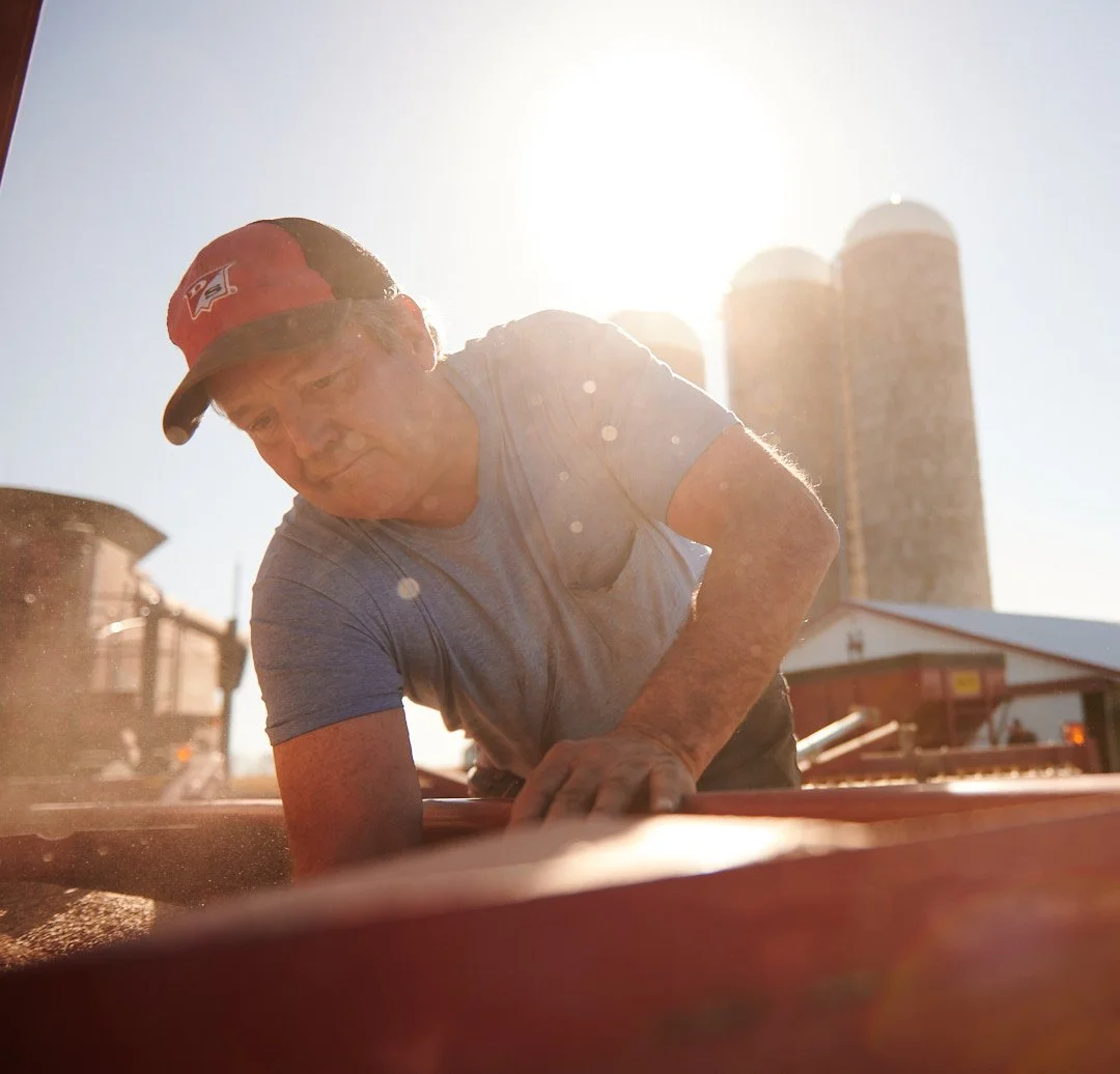 Man in a gray t-shirt and red cap working outdoors on a farm with silos in the background during sunset.