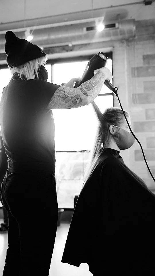 Hairdresser blow-drying a client's hair in a salon with large window and brick wall background