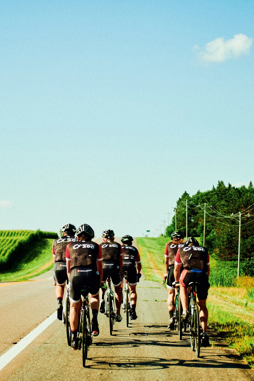 Group of cyclists riding on a rural road surrounded by green fields and trees, under a blue sky with a few clouds.