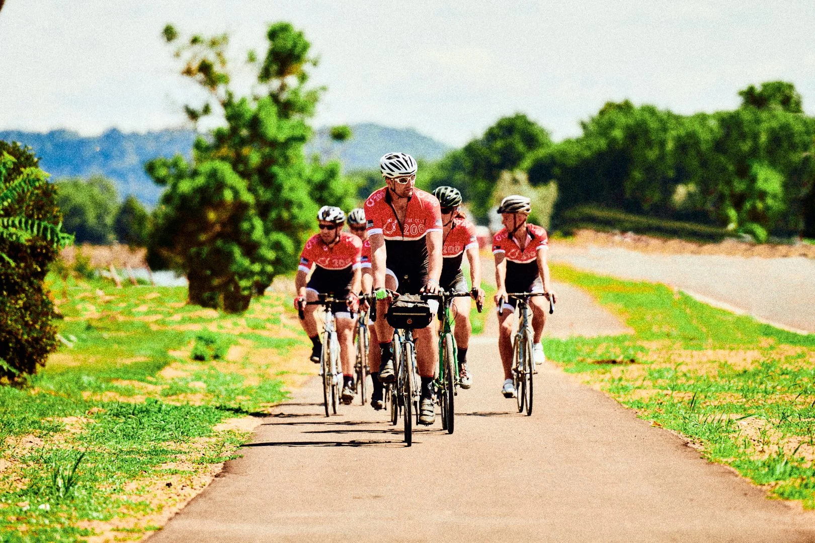 A group of cyclists riding on a paved path through a lush green landscape with trees and bushes, under a partly cloudy sky.