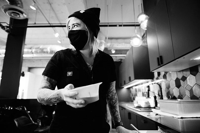 A woman with tattoos wearing a face mask and a beanie, holding a container in a modern restaurant kitchen.