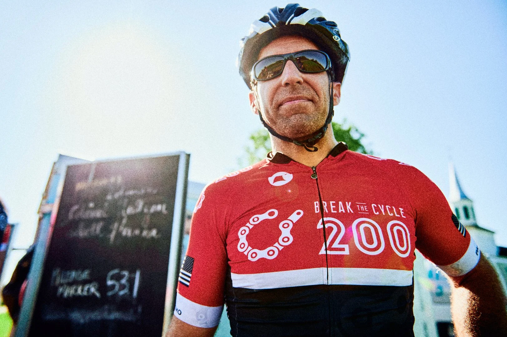A man wearing a red cycling jersey with the text "Break the Cycle 200" and a chain graphic, sunglasses, and a black helmet, standing outdoors on a sunny day.
