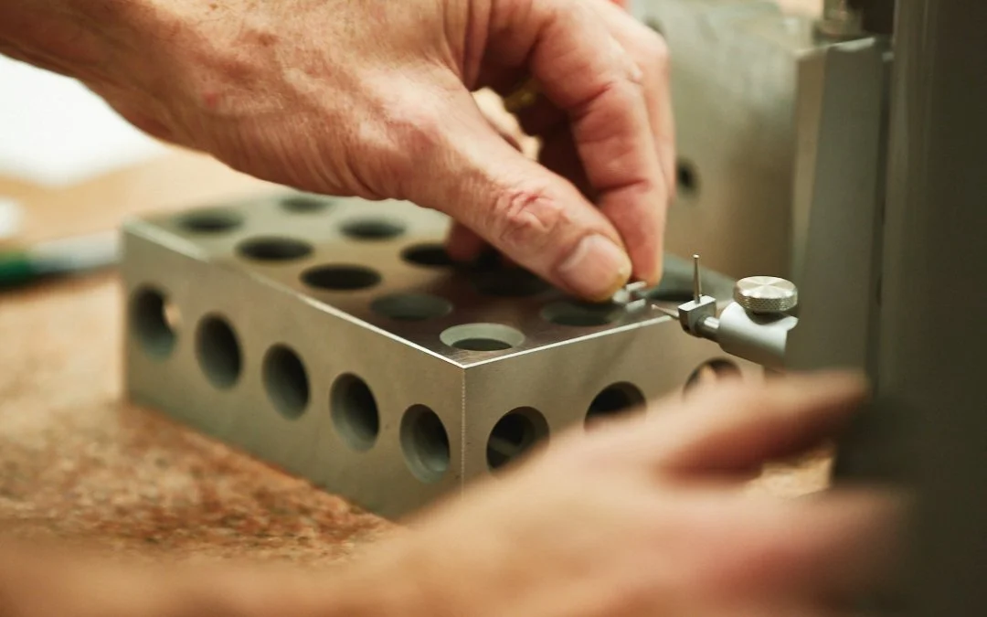 Close-up of a person's hands working on a metal block with multiple holes, possibly in a manufacturing or laboratory setting.