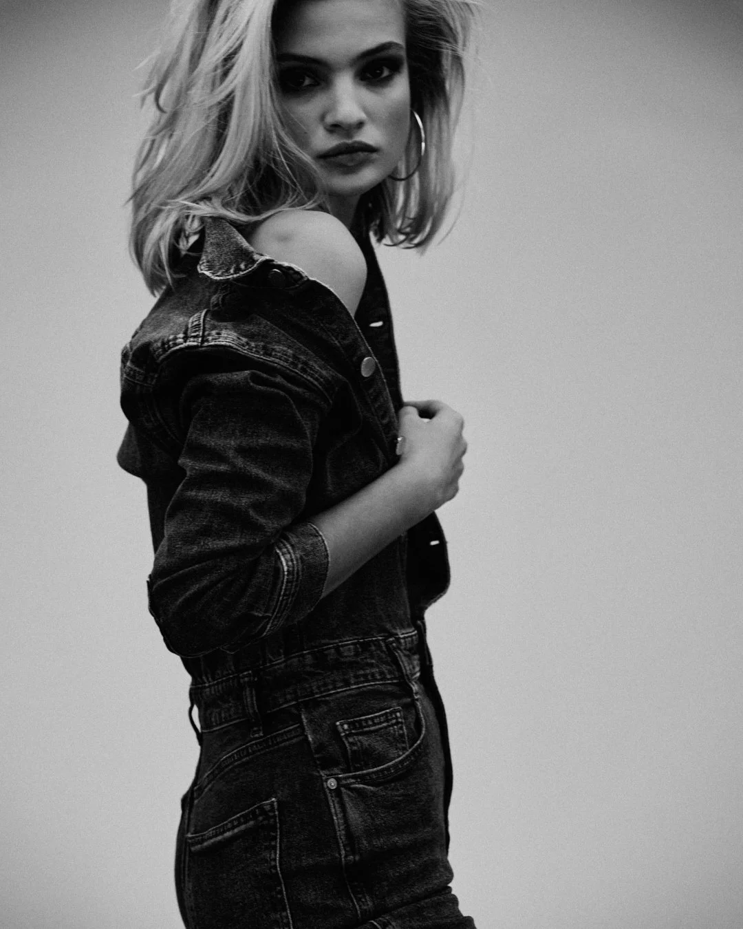 Black and white photo of a young woman with shoulder-length hair wearing a denim jacket off the shoulder, tilting her head slightly to the right, with a serious expression, hoop earrings, and a plain background.