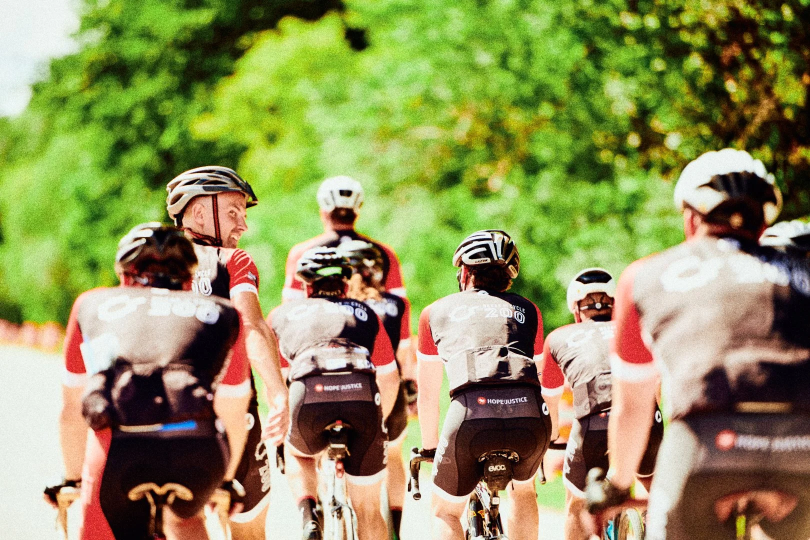 Group of cyclists riding on a sunny trail surrounded by lush green trees, wearing helmets and sports gear.