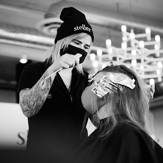 A hairstylist with tattoos, wearing a beanie and face mask, applying hair dye or foil highlights to a client's hair in a salon.