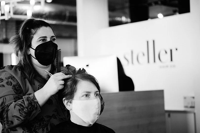 A hairstylist wearing a black face mask trims a woman's hair, who is wearing a white face mask, inside a hair salon.