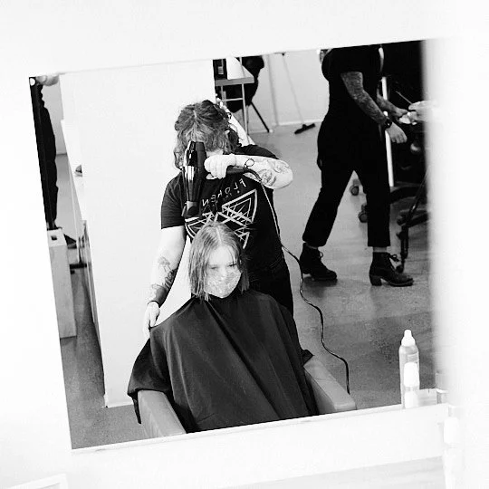 Hairdresser styling a woman's hair in a salon, woman sitting in a salon chair wearing a face mask