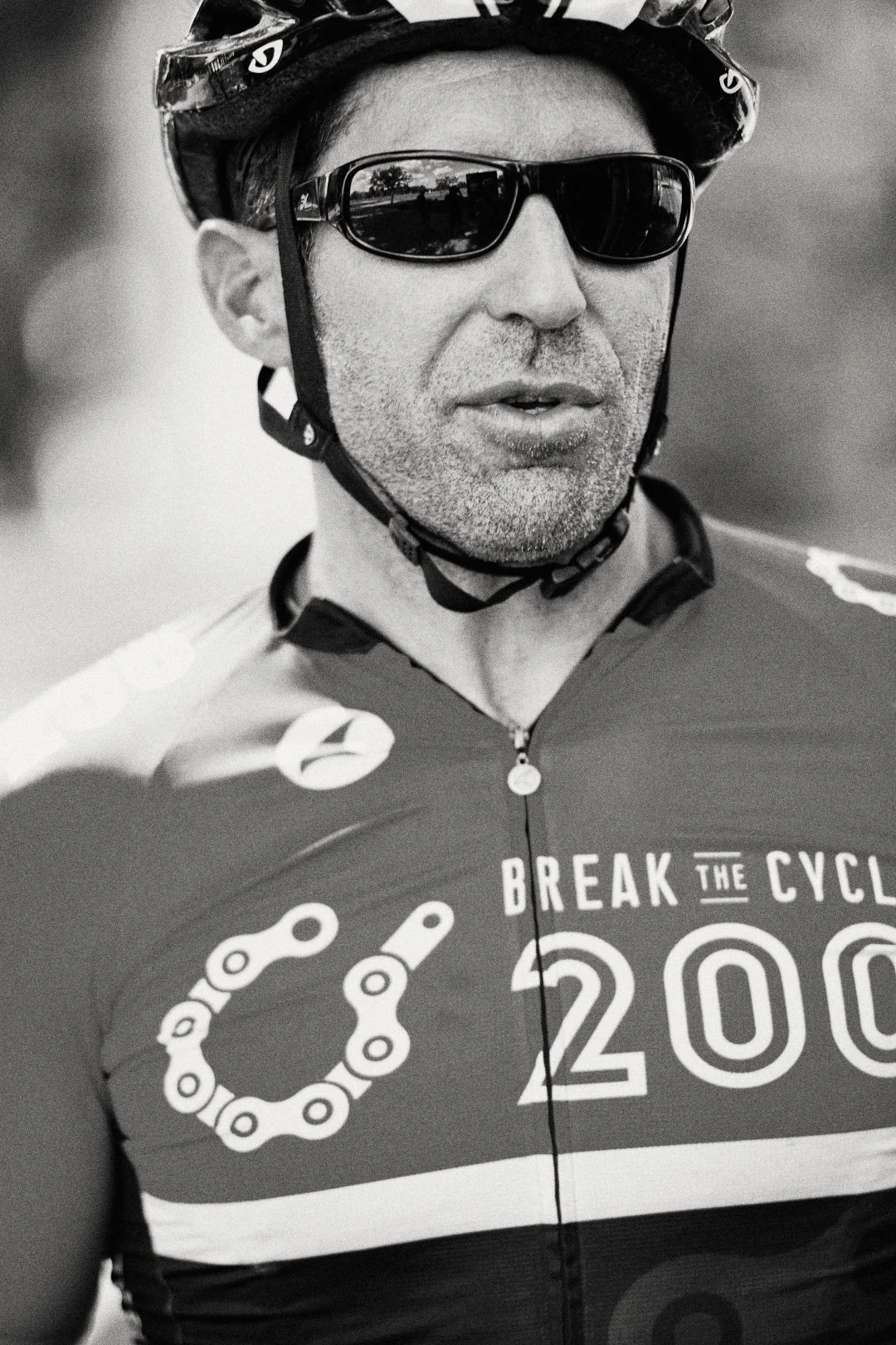 A man wearing sunglasses, a helmet, and cycling gear with the words 'Break the Cycle 200'. he appears to be participating in a cycling event.