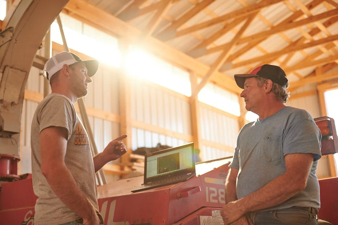 Two men in a barn, engaged in conversation, with a laptop on a red farm equipment. One man is wearing a beige t-shirt and cap, and the other is in a gray t-shirt and baseball cap. Bright sunlight streams in through the barn's open windows.