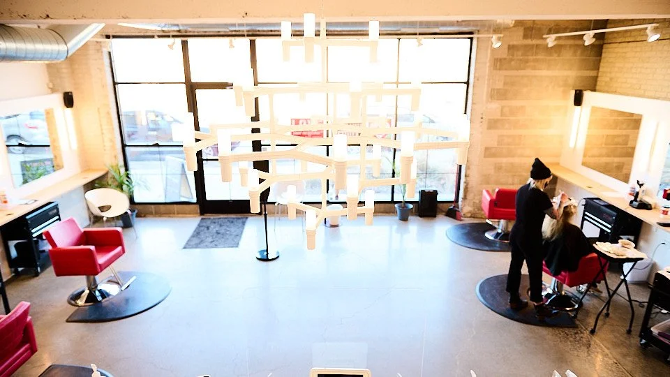 A hair salon with large windows, red salon chairs, and a stylist working on a client. Modern lighting fixture hangs from the ceiling, and there are mirrors and work stations along the walls.
