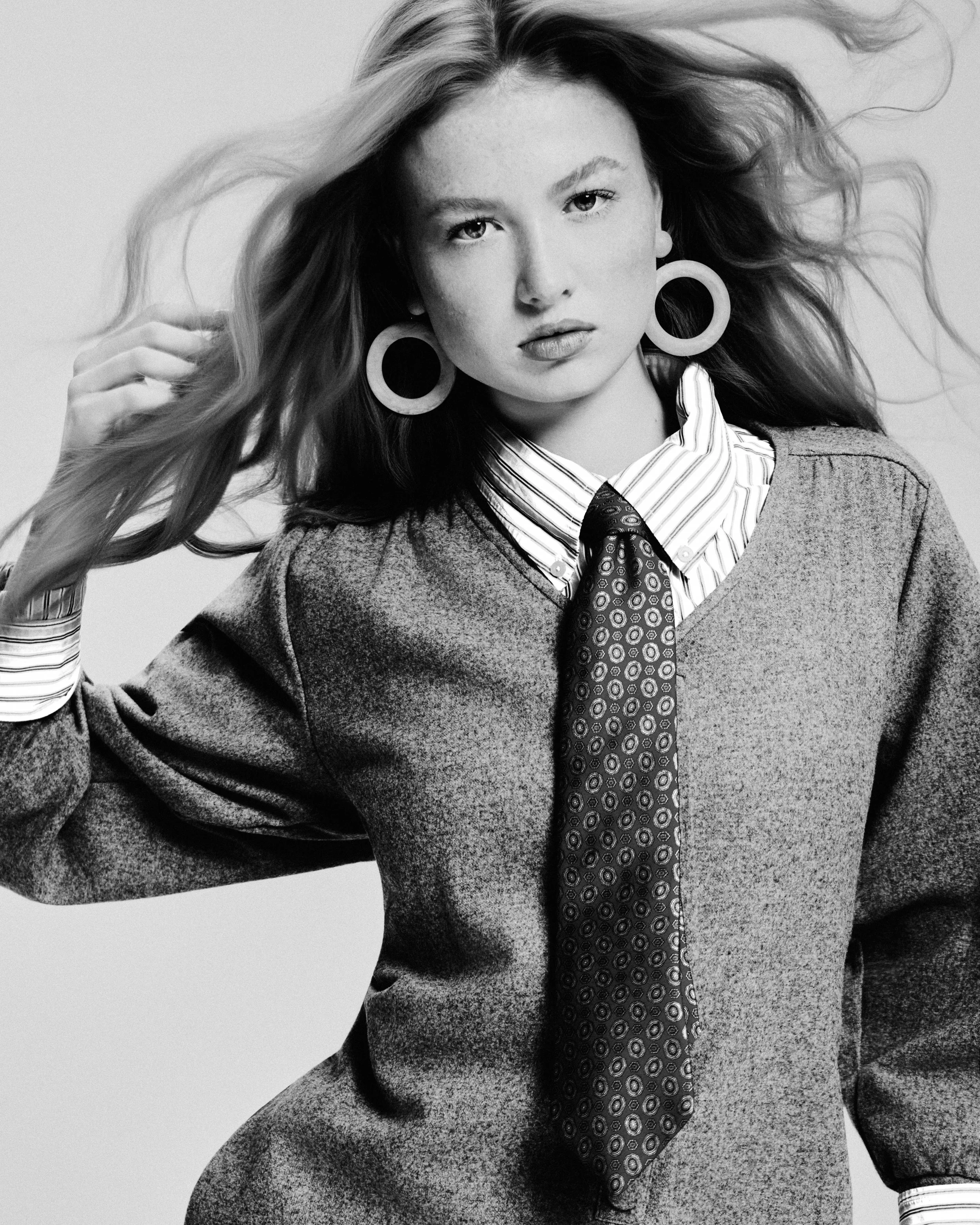 Black and white portrait of a woman with long hair, wearing large circular earrings, a striped shirt, a tie, and a sweater.