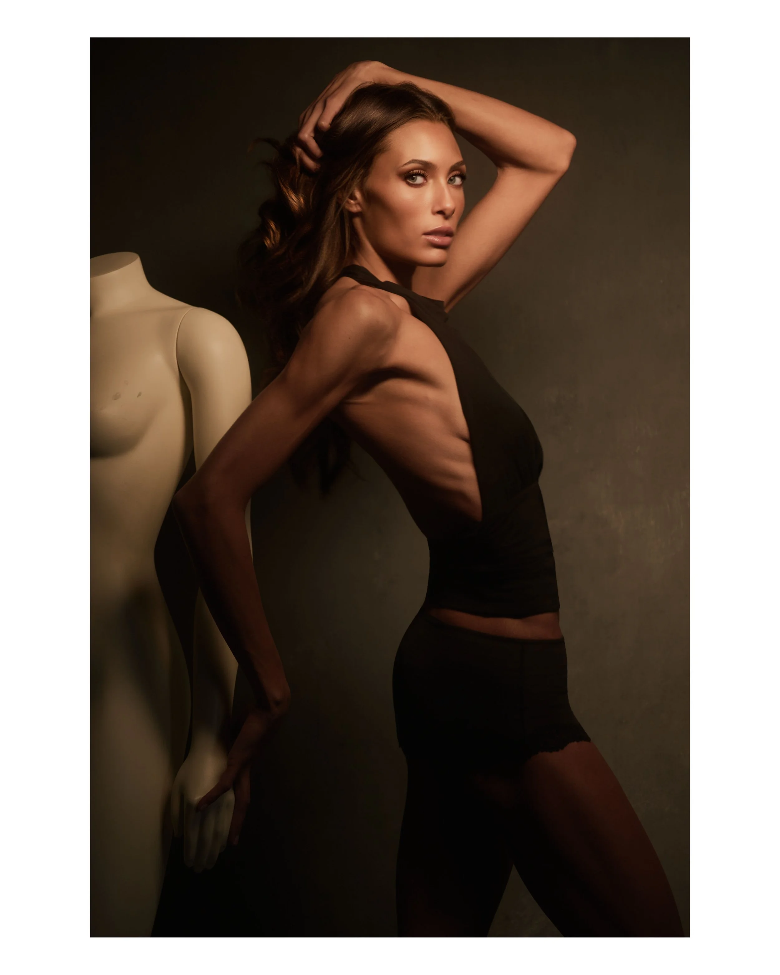 A woman with long brown hair posing next to a torso mannequin in a studio with dark background, wearing a black sleeveless outfit.