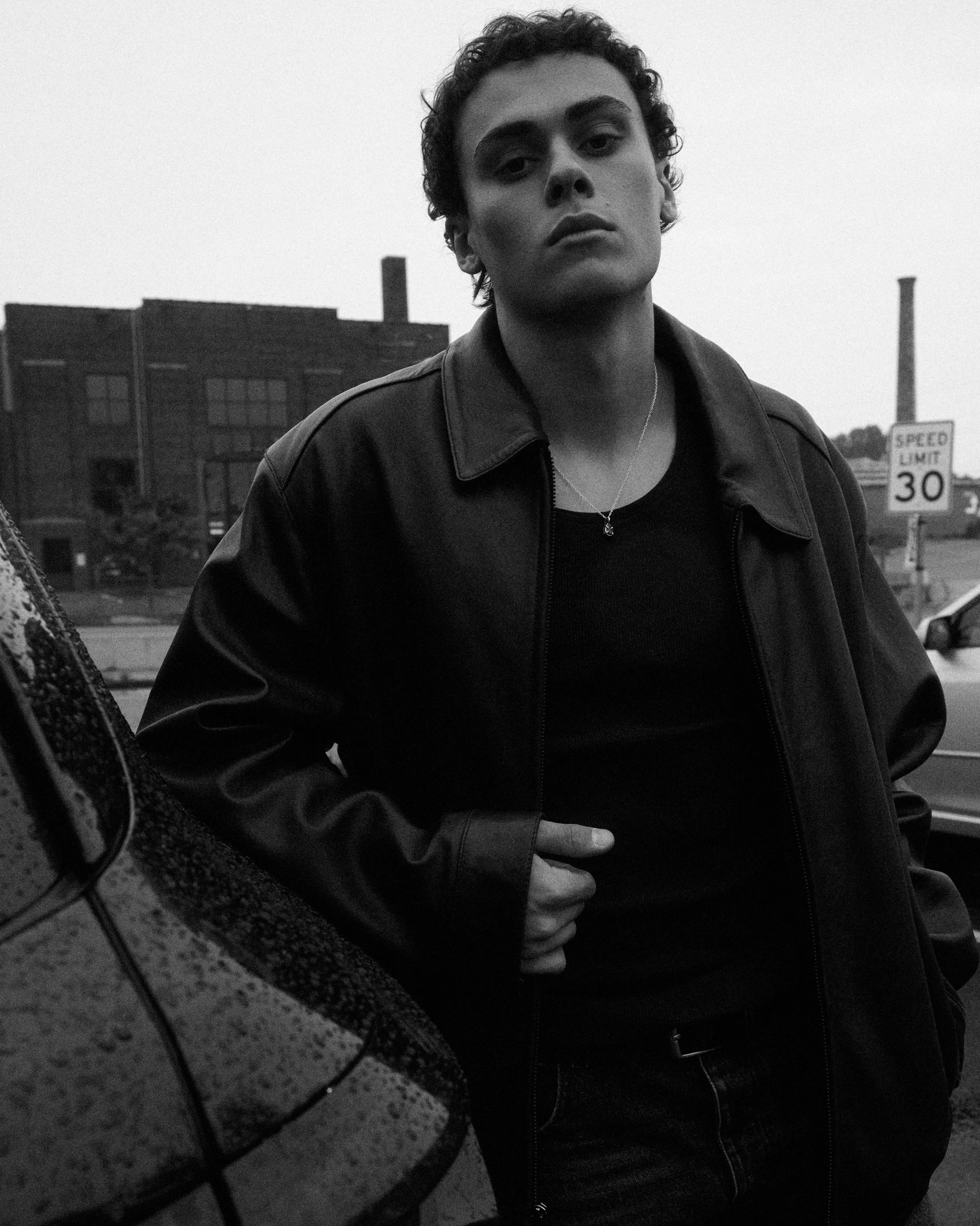 A black and white photo of a young man with curly hair leaning against a car, wearing a leather jacket and a necklace, with an industrial background and a speed limit sign.