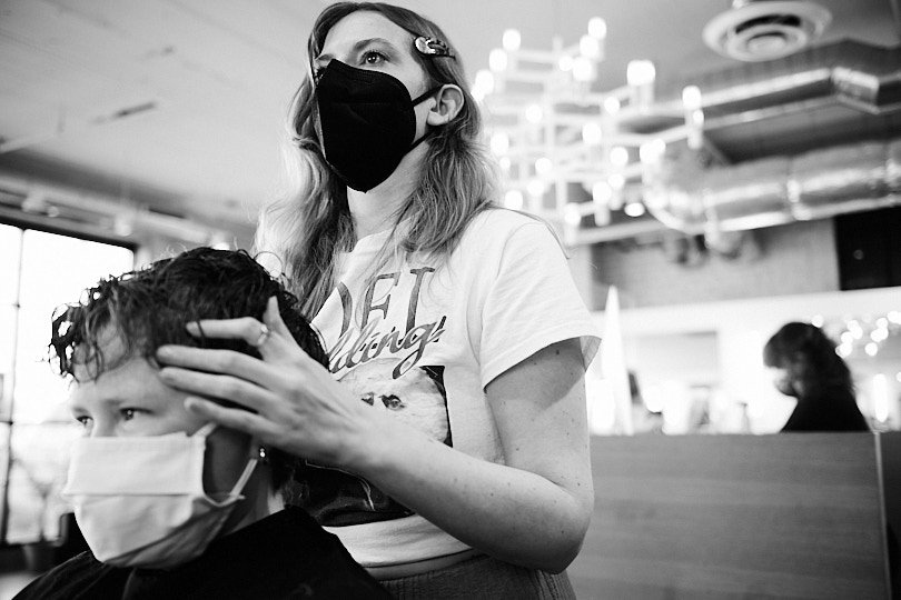 A woman wearing a black face mask and casual t-shirt giving a haircut to a person with curly hair, who is wearing a white face mask, in a hair salon.