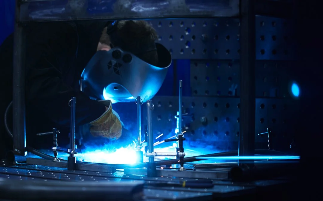 Person welding metal with bright blue sparks in a workshop, wearing protective gear.