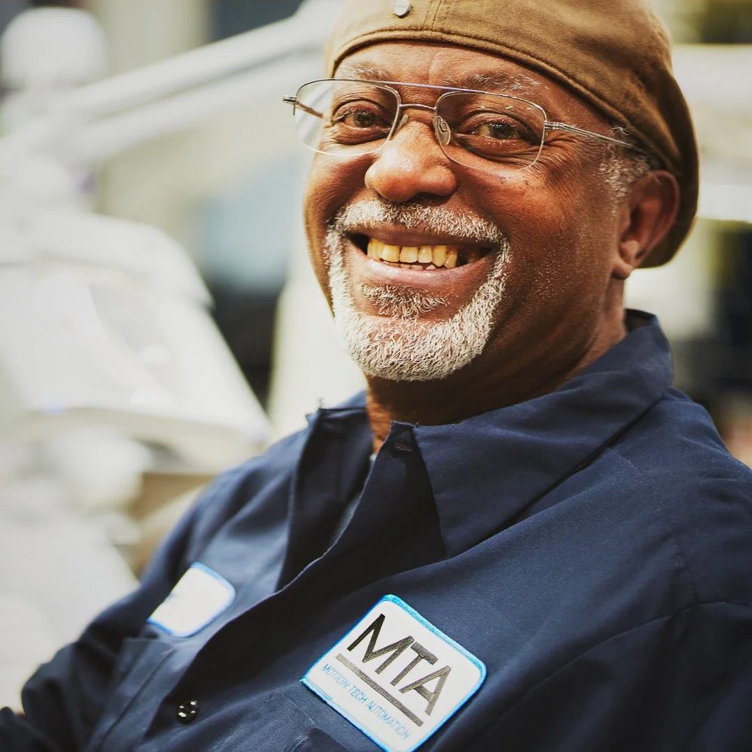 Portrait of a smiling older man with glasses, wearing a brown cap and a navy uniform with an 'ATM' patch, outdoors in bright lighting.