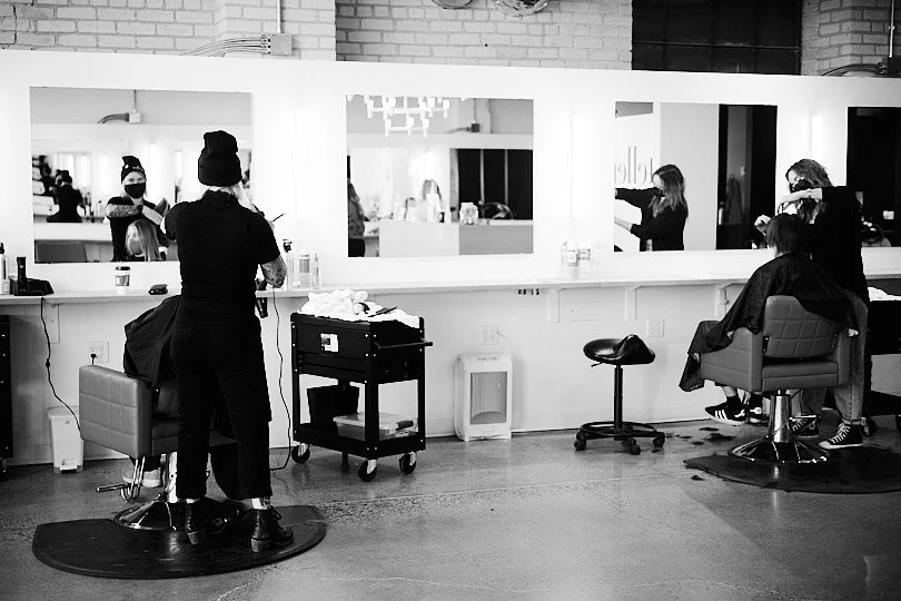 Black and white photo of a barbershop with three hairstylists working on clients. Mirrors line the wall behind them, and various hair styling tools are visible.