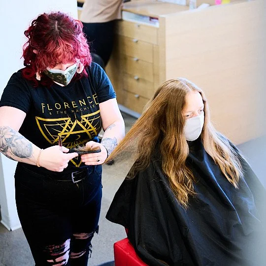A hairdresser with pink hair and tattoos is styling a woman's long red hair at a salon. Both are wearing face masks.