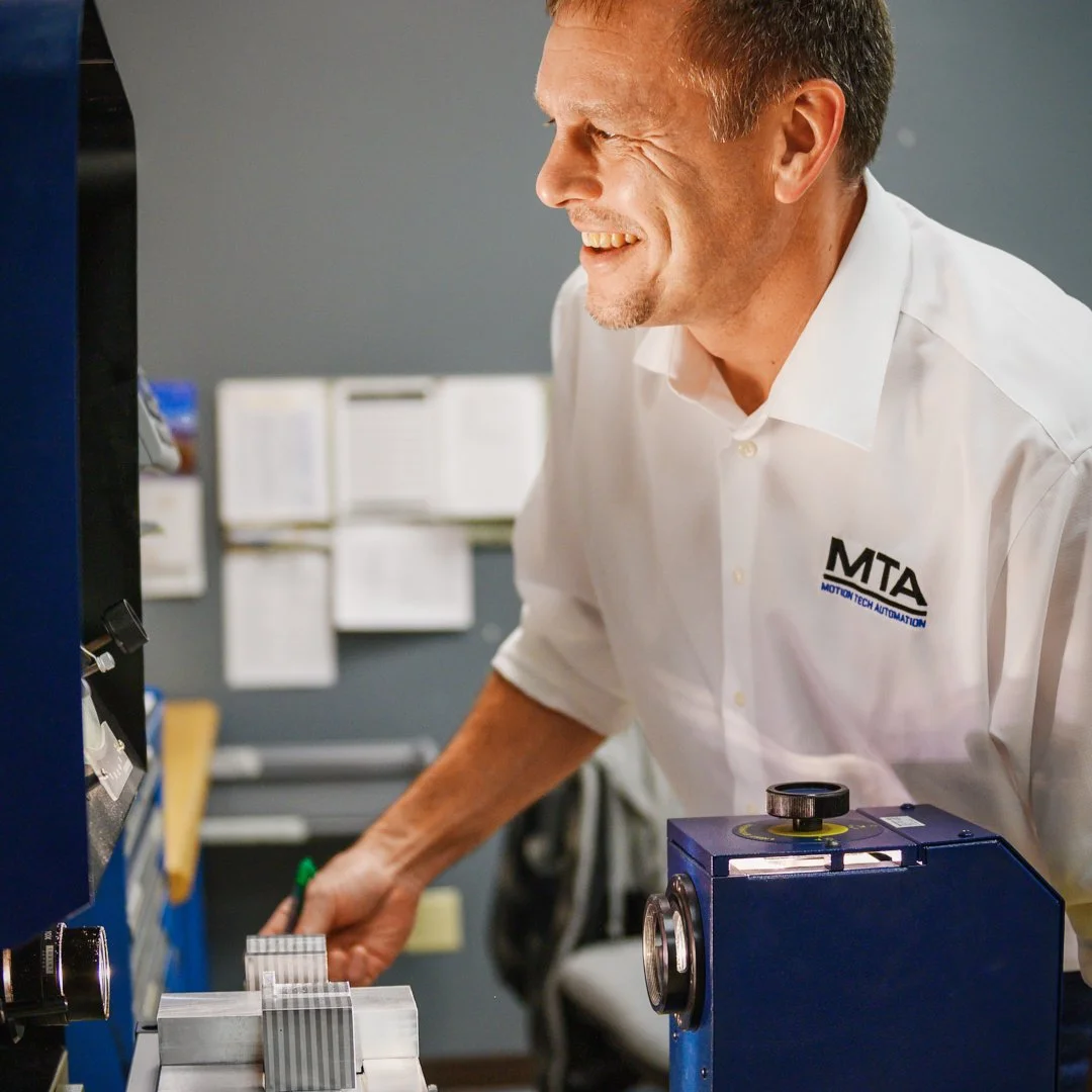 A smiling man in a white shirt with 'MTA' logo and 'Motion Tech Automation' text working with machinery in an industrial setting.