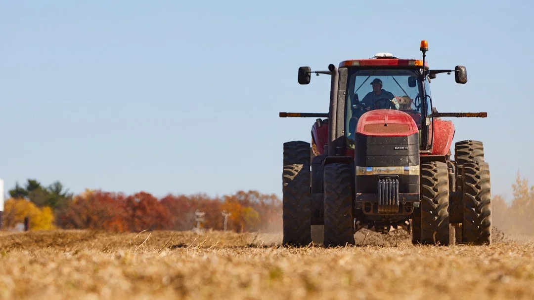 Red tractor working on a farm field in fall with trees showing autumn colors in the background