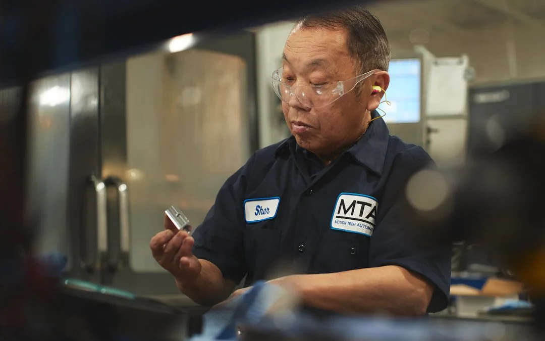 A mechanic wearing safety glasses and a navy blue work shirt with name tag 'Sher' and 'MTA' patch, working in an auto repair shop.