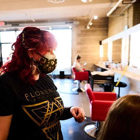 A hairstylist wearing a black T-shirt with a geometric design and a camouflage mask, working on a client's hair in a modern salon.