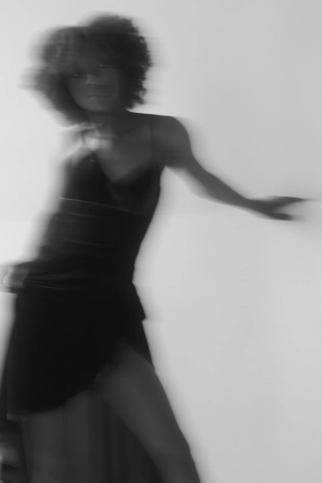 A blurred black and white photo of a woman with curly hair wearing a sleeveless dress, standing against a plain background.
