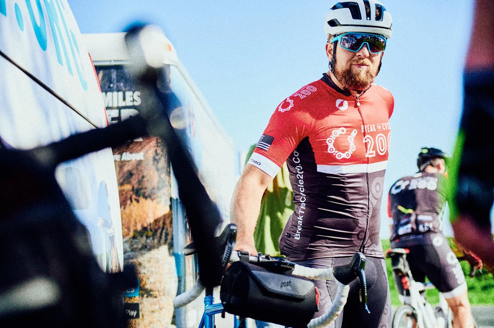 A man wearing a red and black cycling jersey with the number 200 and a chain design, standing outdoors with his bike, wearing a white helmet and sunglasses. Another cyclist is visible in the background.