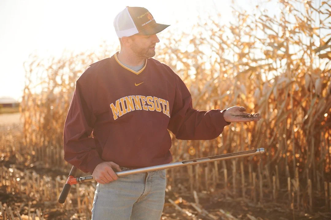 A man wearing a Minnesota sweatshirt and baseball cap standing in a cornfield at sunset, holding a muddy hand and a gardening tool.