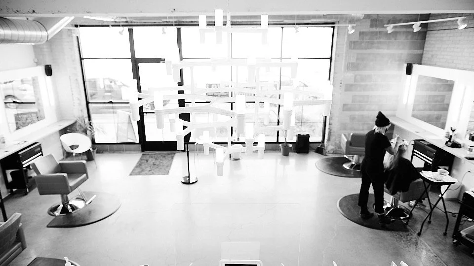 A hair salon with large windows, modern lighting fixtures, and two stylists working on clients, with salon chairs and mirrors.