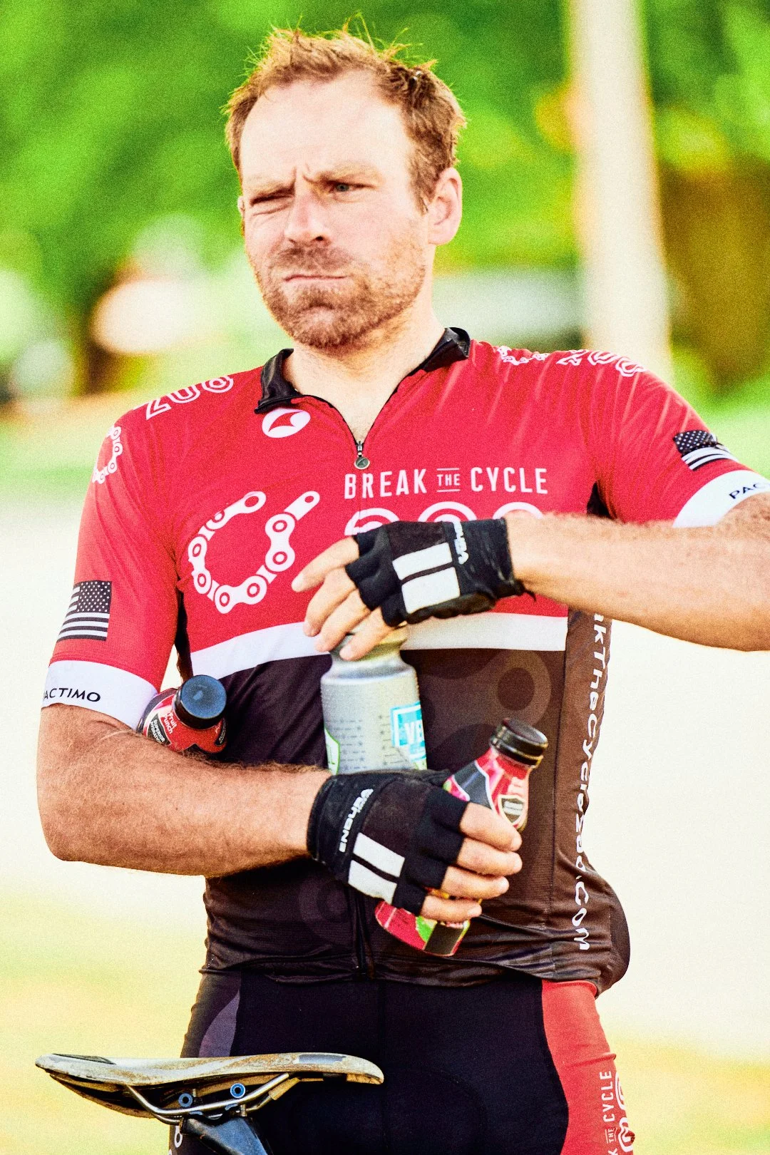 A man in cycling gear with a red jersey, standing outdoors with a green, blurred background, holding water bottles.